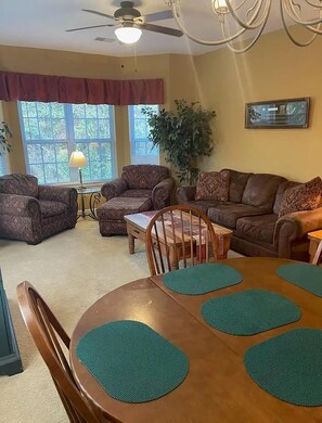 A living room with a dining table, two sofas, a ceiling fan, and a large window with blinds. - Pool Days & Golf Getaways in Myrtle Beach (Myrtle Beach)