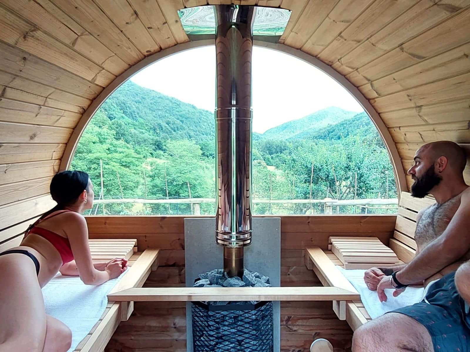 Una baita di legno con un'ampia finestra ad arco che si affaccia su una montagna ricoperta di bosco. All'interno, si trovano panche di legno e un sistema di riscaldamento centralizzato.