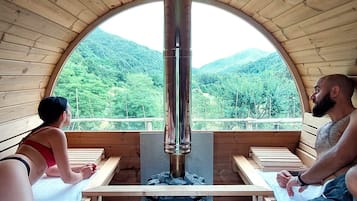 Una baita di legno con un'ampia finestra ad arco che si affaccia su una montagna ricoperta di bosco. All'interno, si trovano panche di legno e un sistema di riscaldamento centralizzato.