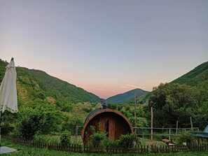 A wooden barrel structure with a door, surrounded by a wooden fence, set in a lush green valley with hills in the background. - Wellness Retreat in the Countryside (Varese Ligure)