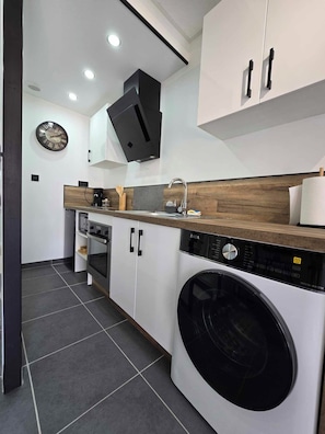 A modern kitchen with a built-in washing machine, a clock on the wall, and a wooden countertop. - New apartment near the Outlet Village (La Verpillière)