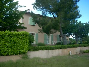 Un bâtiment à deux étages avec une façade beige, des volets verts et un grand arbre devant.