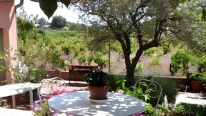 Un patio exterior con una mesa redonda y sillas, rodeado de plantas en macetas y vistas a un jardín ajardinado.