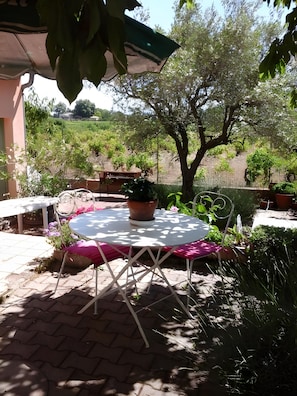 Une terrasse extérieure avec une table ronde et des chaises, entourée de plantes en pot et offrant une vue sur un jardin aménagé.