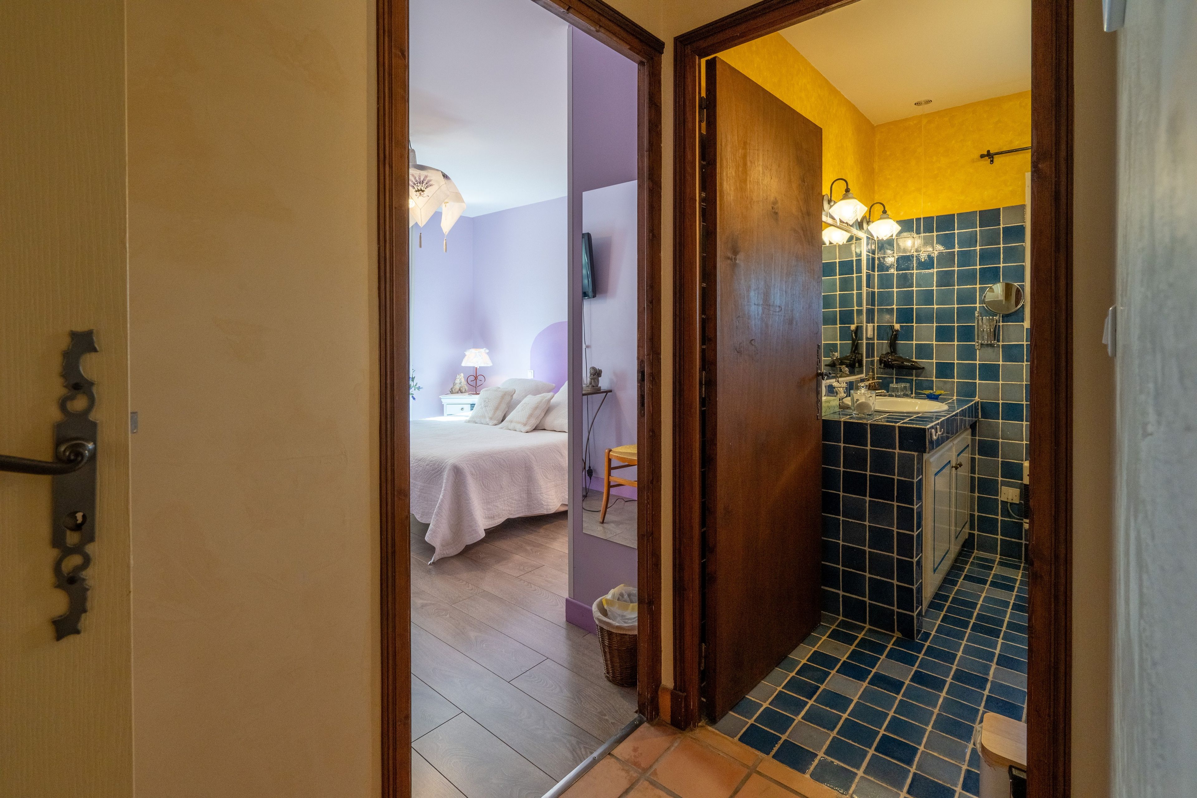 Una habitación con una cama, una mesita de noche, una lámpara y un baño con lavamanos y espejo.