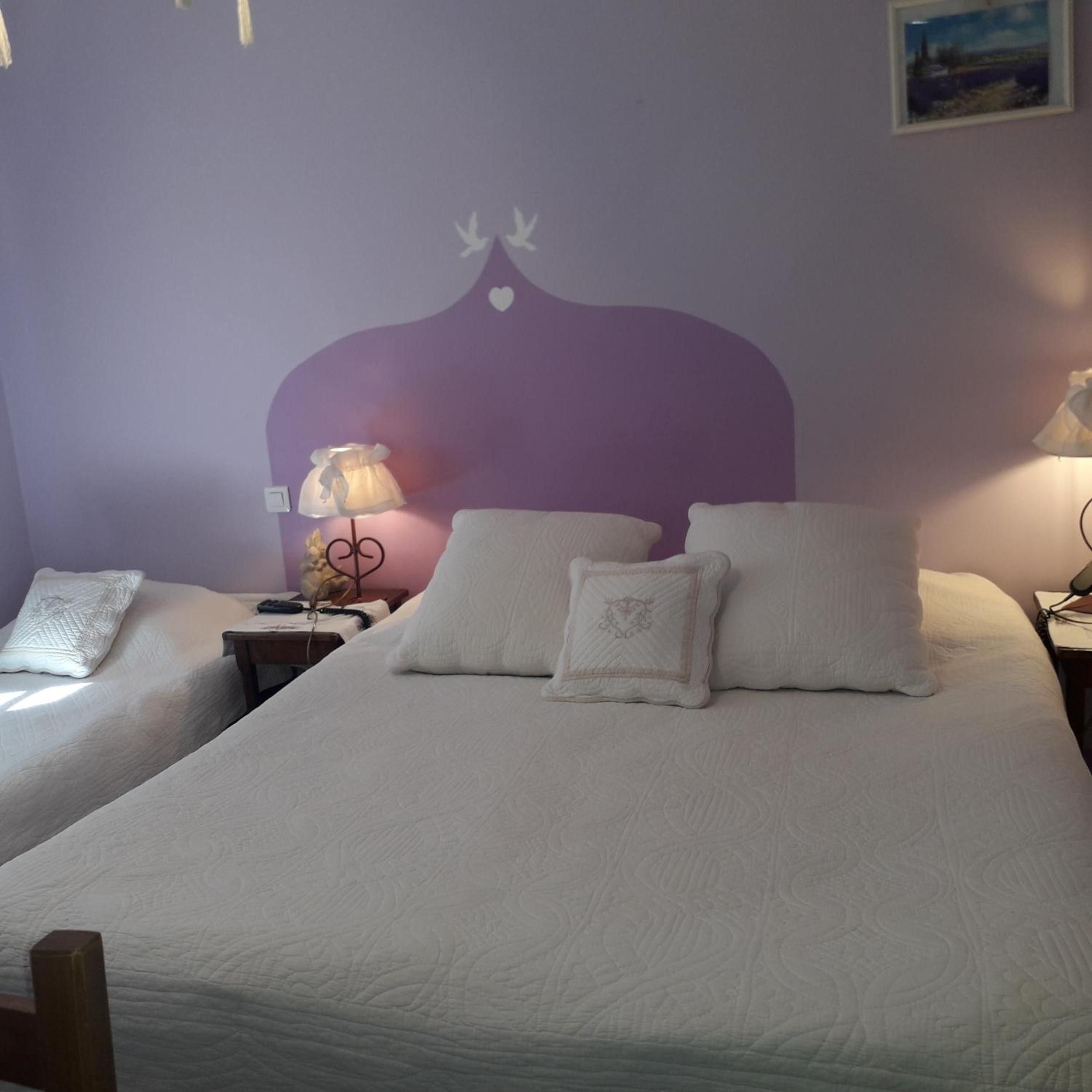 Ein Doppelzimmer mit zwei Betten, jeweils mit weißer Bettwäsche und Kissen, einer violetten Akzentwand und einem kleinen Tisch mit einer Lampe.
