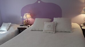 Habitación doble con dos camas, cada una con ropa blanca de cama y almohadas, una pared de acento morada y una mesita con una lámpara.