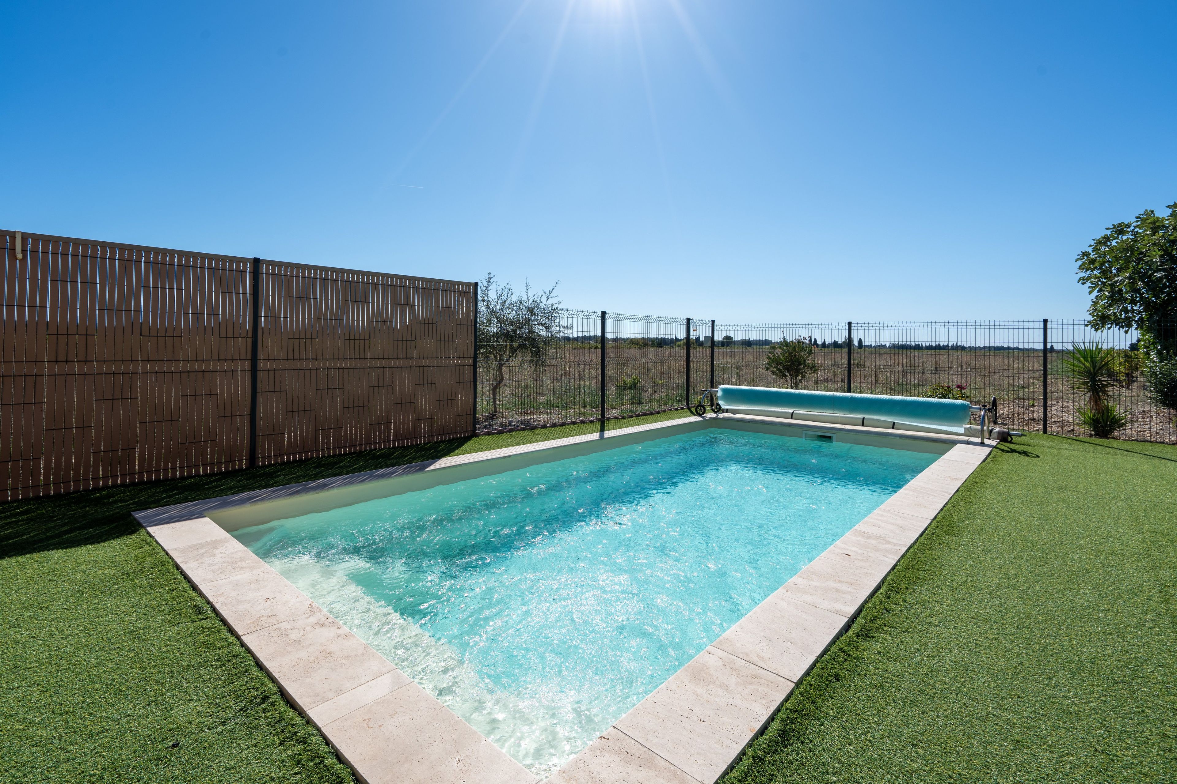 A rectangular swimming pool with a clear blue water surface, surrounded by a low hedge and a tall fence.
