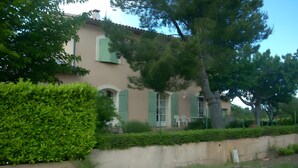 Un edificio de dos plantas con fachada color beige, contraventanas verdes y un gran árbol en la parte delantera.