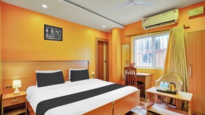 Een hotelkamer met een bed, nachtkastje, lamp, airconditioning, raam met gordijnen en een kleintje tafeltje met een stoel.