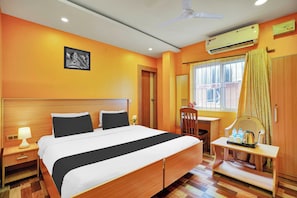 Habitación de hotel con cama, mesita de noche, lámpara, aire acondicionado, ventana con cortinas y una pequeña mesa con una silla.
