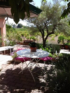 Une terrasse extérieure avec une table ronde et des chaises, entourée de plantes en pot et offrant une vue sur un jardin aménagé.