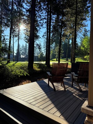 Een terras met houten vloer, twee Adirondack stoelen en uitzicht op een bos met hoge bomen en een heldere lucht.