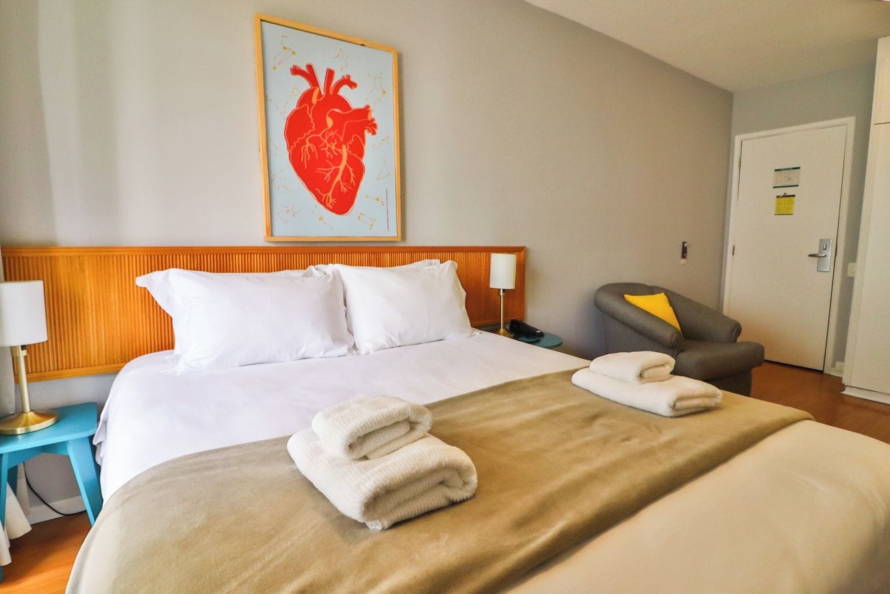 Quarto de hotel com cama, mesa de cabeceira com abajur, uma cadeira e porta.