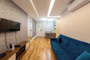 Uma sala de estar moderna com televisão de tela plana, piso de madeira, um sofá azul e uma área de jantar com mesa e cadeiras.