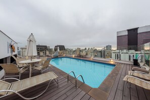 Piscina no terraço com espreguiçadeiras e guarda-sóis, em meio a edifícios da cidade.