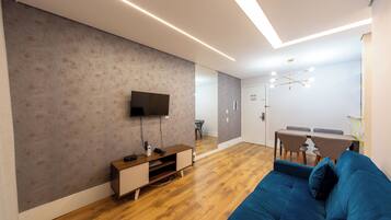 Uma sala de estar moderna com televisão de tela plana, piso de madeira, um sofá azul e uma área de jantar com mesa e cadeiras.