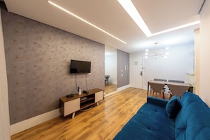 Uma sala de estar moderna com televisão de tela plana, piso de madeira, um sofá azul e uma área de jantar com mesa e cadeiras.