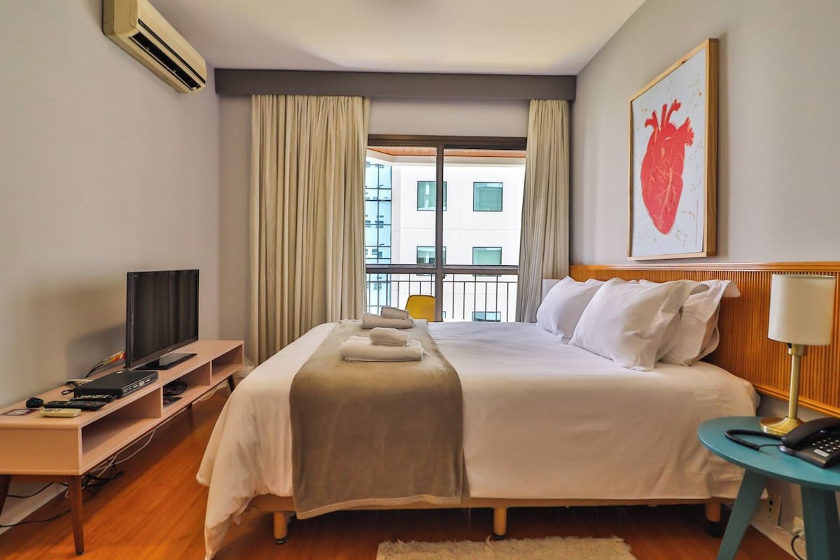 Quarto de hotel com cama, televisão, escrivaninha, cadeira, janela com cortinas e obra de arte na parede.