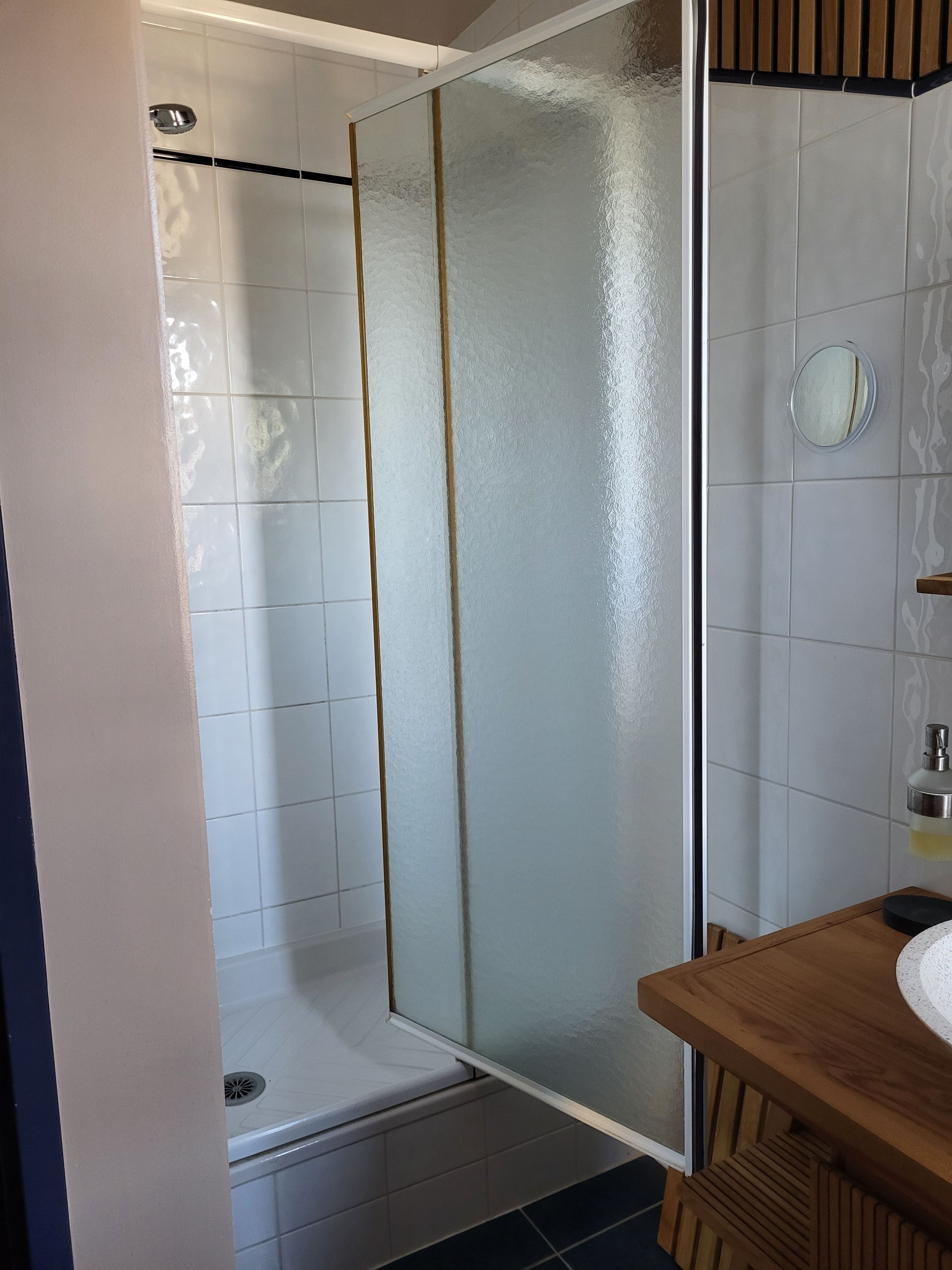 Een badkamer met een douchecabine van glas, witte tegels, een houten wastafelmeubel en een ronde spiegel.
