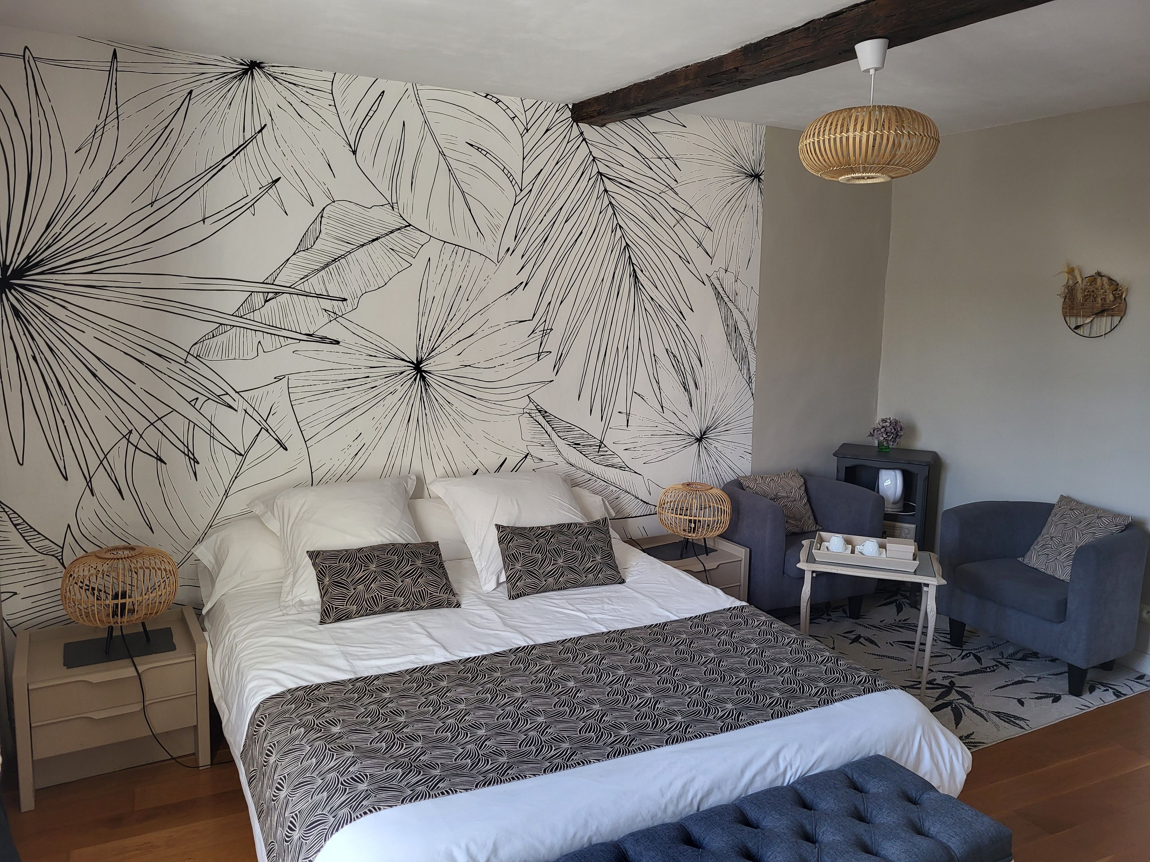 Ein Schlafzimmer mit einem großen Bett, einem Nachttisch, einem Sessel, einem kleinen Tisch mit Lampe und einer Wand mit Blätermotiv.