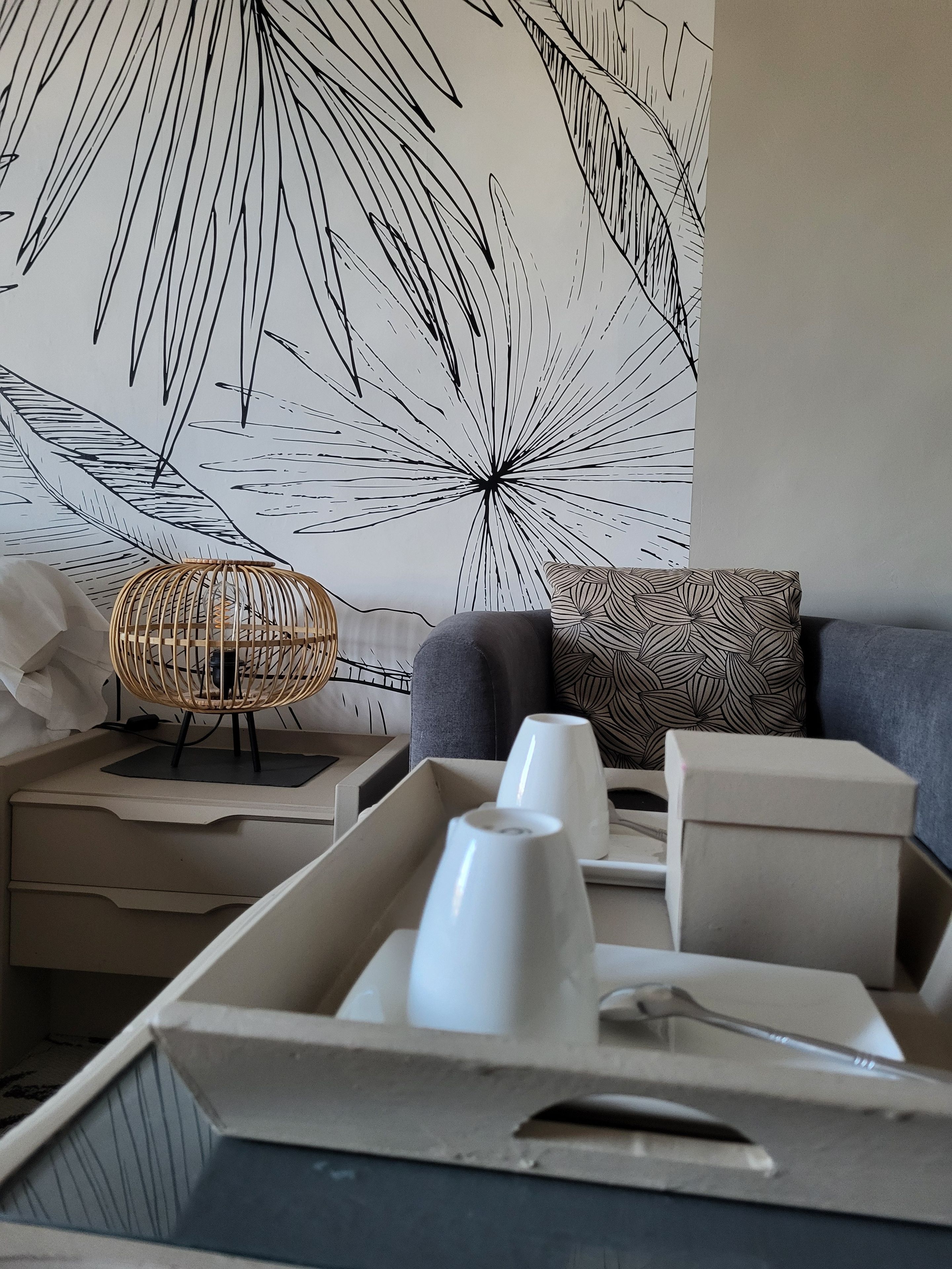 Ein modernes Wohnzimmer mit einer großen Wandmalerei, einem grauen Sofa, einem gemusterten Kissen, einem Korbtisch und einer Platte mit weißen Tassen.