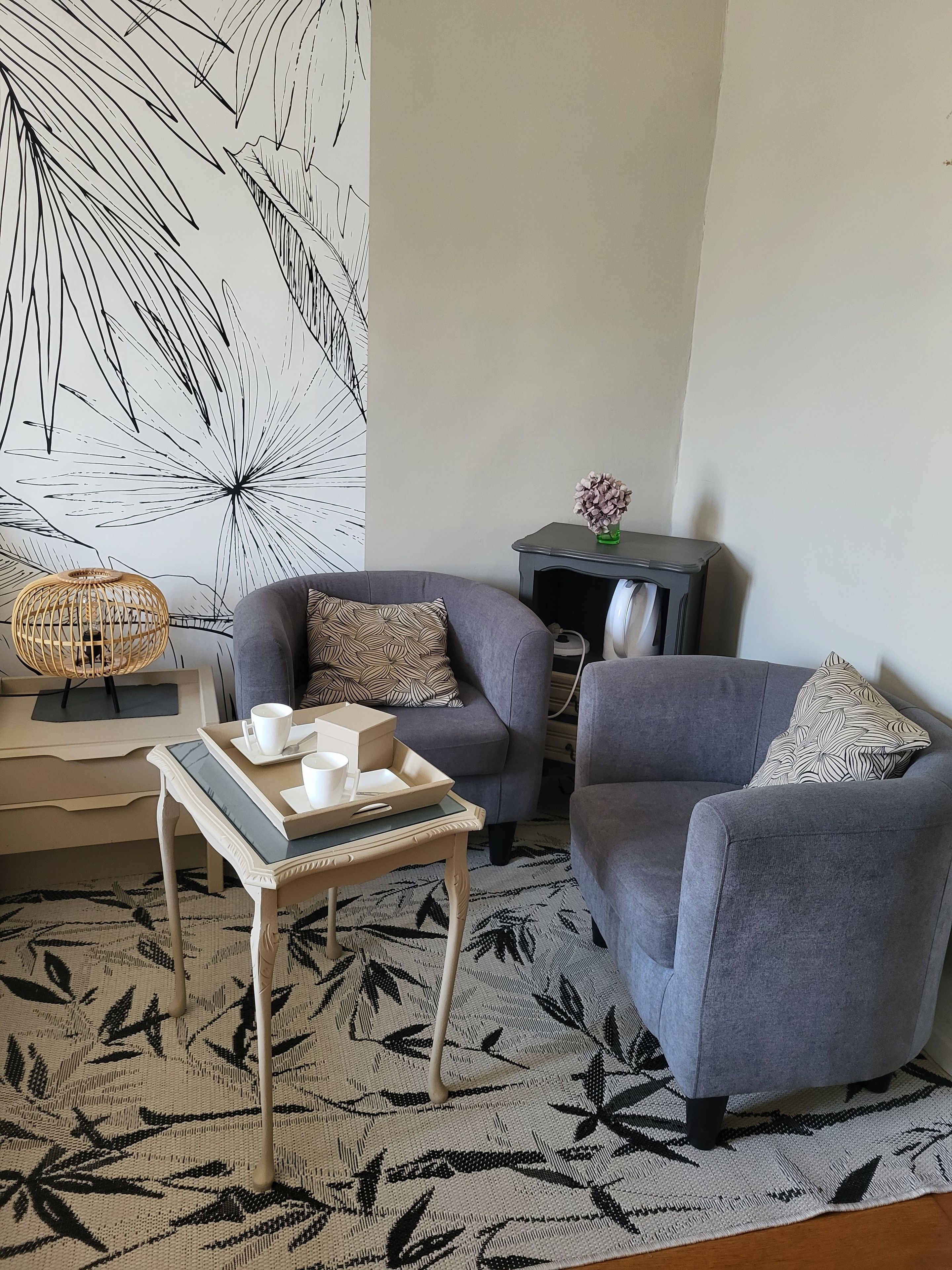 Un salon avec un canapé gris, un tapis à motifs, une petite table d’appoint agrémentée d’un objet décoratif et un mur orné d’un motif abstrait en noir et blanc.