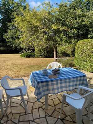 Ein Tisch im Freien mit kariertem Tischläufer, zwei Stühlen und einer Kanne auf einer mit Steinplatten belegten Terrasse, umgeben von Grünpflanzen.