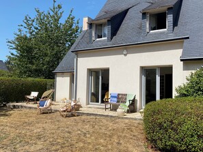 Ein zweigeschossiges Haus mit geneigtem Dach, einer überdachten Terrasse mit Gartenmöbeln und einem gepflegten Rasen.