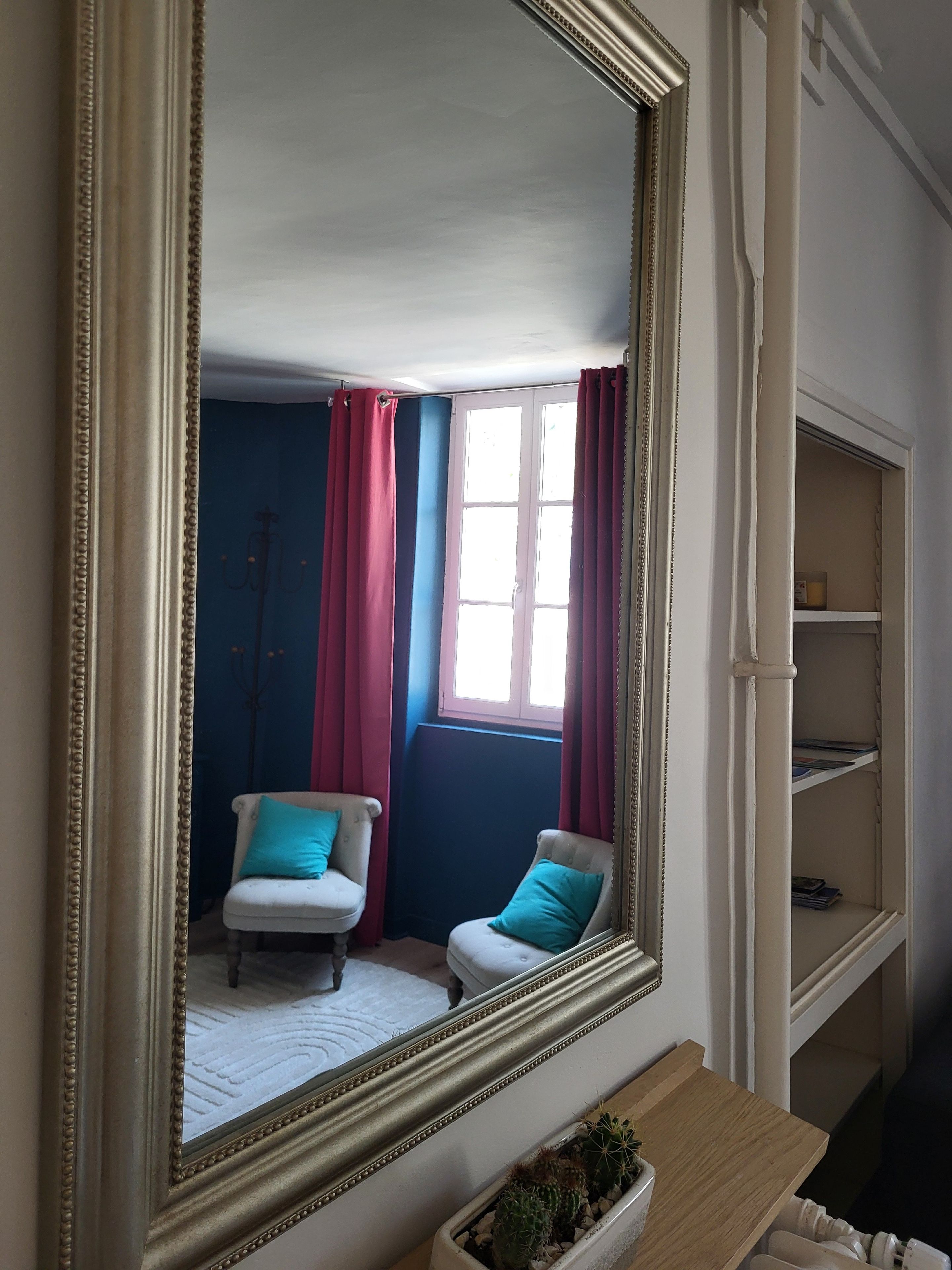 Une pièce avec un grand miroir, une fenêtre ornée de rideaux rouges, un fauteuil blanc et une petite plante posée sur une table en bois.
