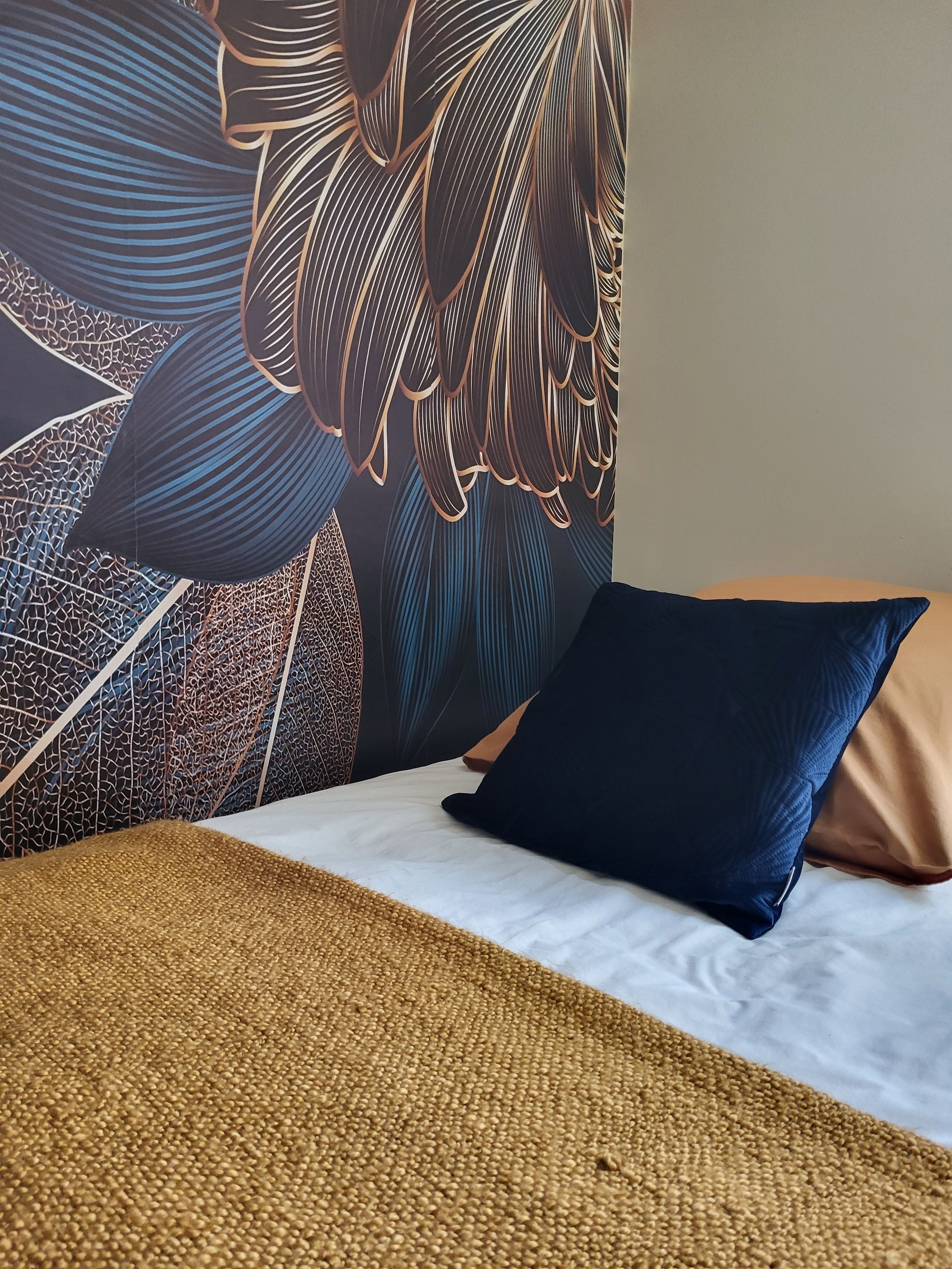Un lit avec une couverture à motifs, un oreiller foncé et un mur orné d’une fresque florale détaillée.
