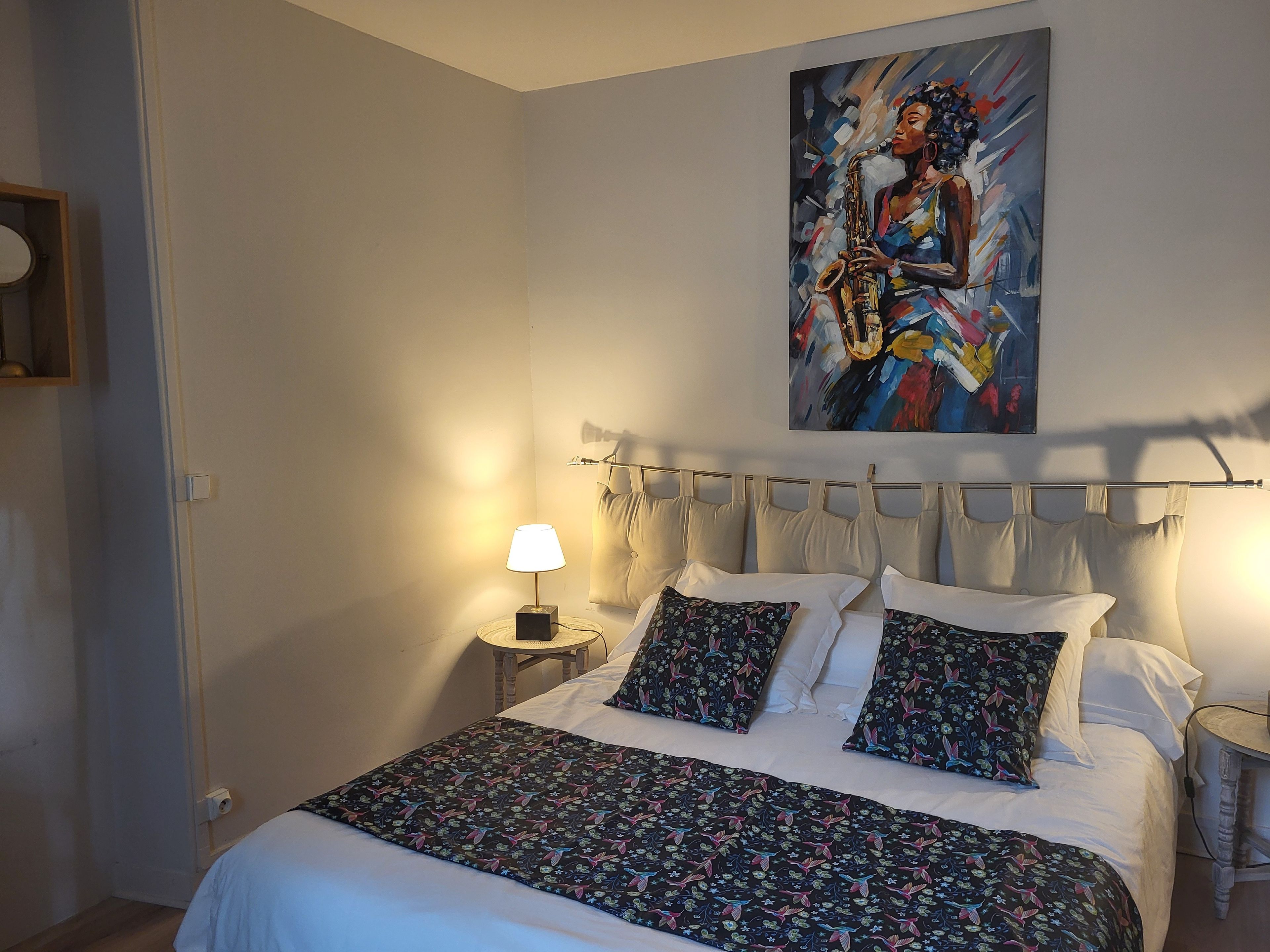 A bedroom with a bed, pillows, a nightstand with a lamp, and a painting on the wall.
