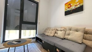 Olohuoneessa on harmaa sohva, pieni pyöreä pöytä, jonka päällä on kaukosäädin, ja seinällä on taulu.