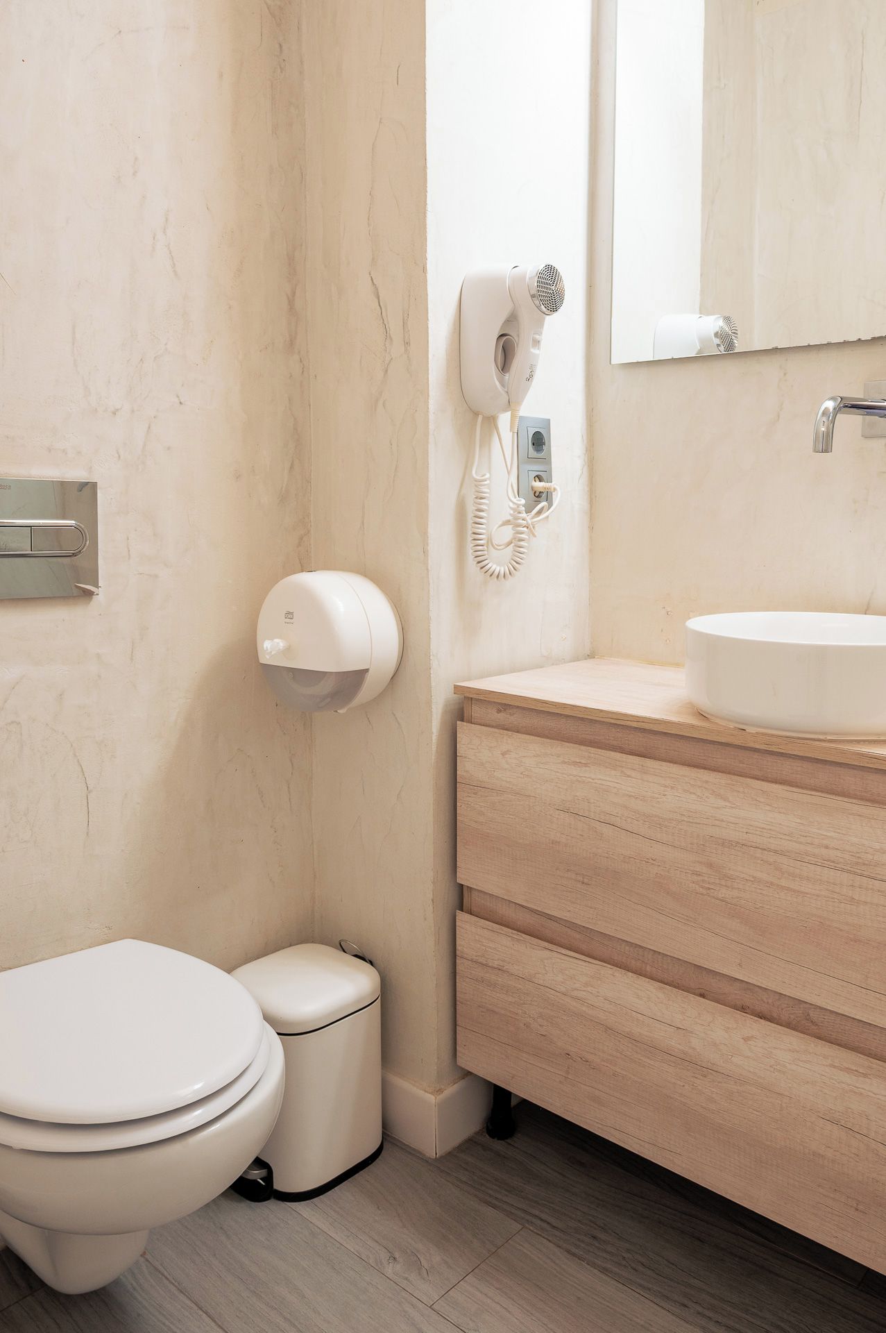Un baño con inodoro, un cubo de basura, un secador de pelo montado en la pared, un secamanos y un tocador de madera con lavamanos.