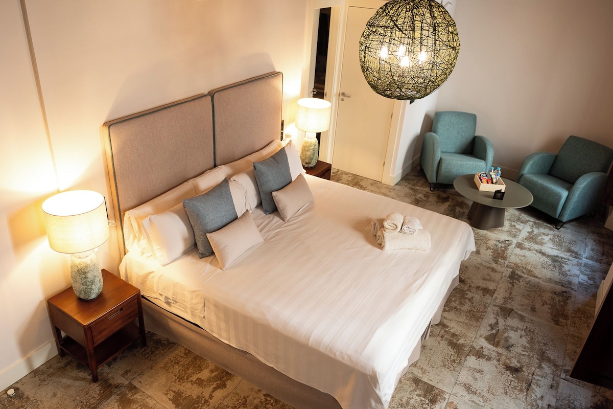 Ein Hotelzimmer mit einem großen Bett, zwei Nachttischlampen, einer Pendelleuchte, zwei Sesseln und einem kleinen Tisch mit Toilettenartikeln.