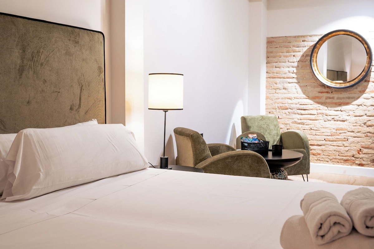 Ein Hotelzimmer mit Bett, Sessel, Tisch, Lampe und rundem Spiegel an einer Ziegelwand.