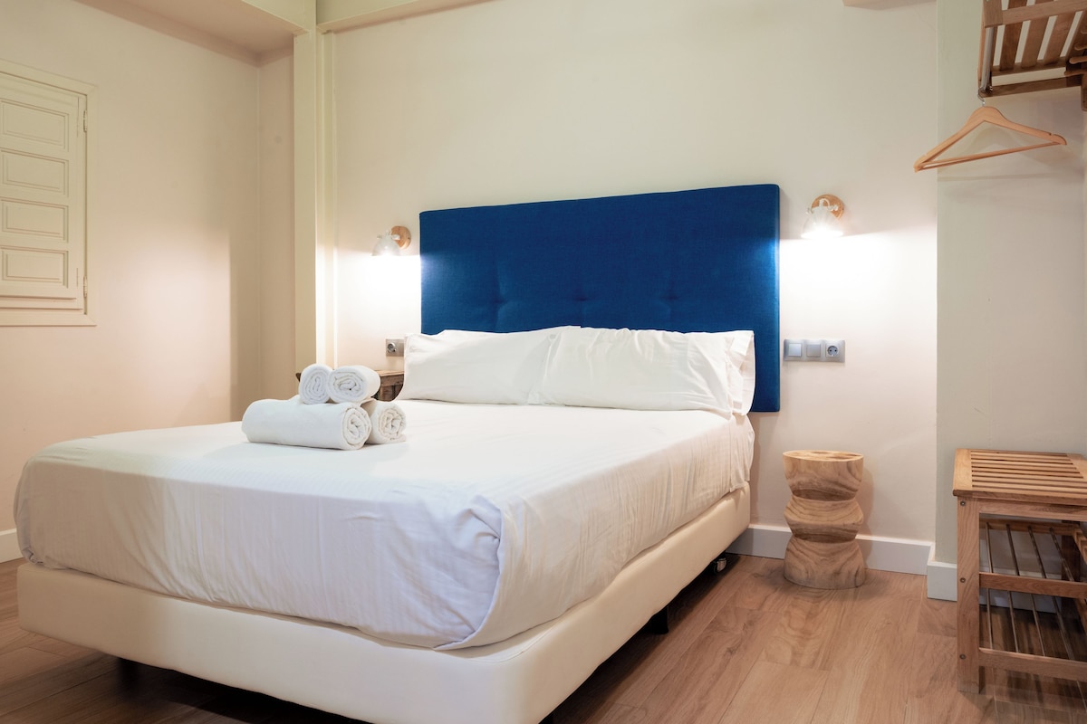 Ein ordentlich gemachtes Bett mit weißen Leinen und zusammengerollten Handtüchern, ein blauer Kopfteil und ein Nachttisch mit einer Lampe.