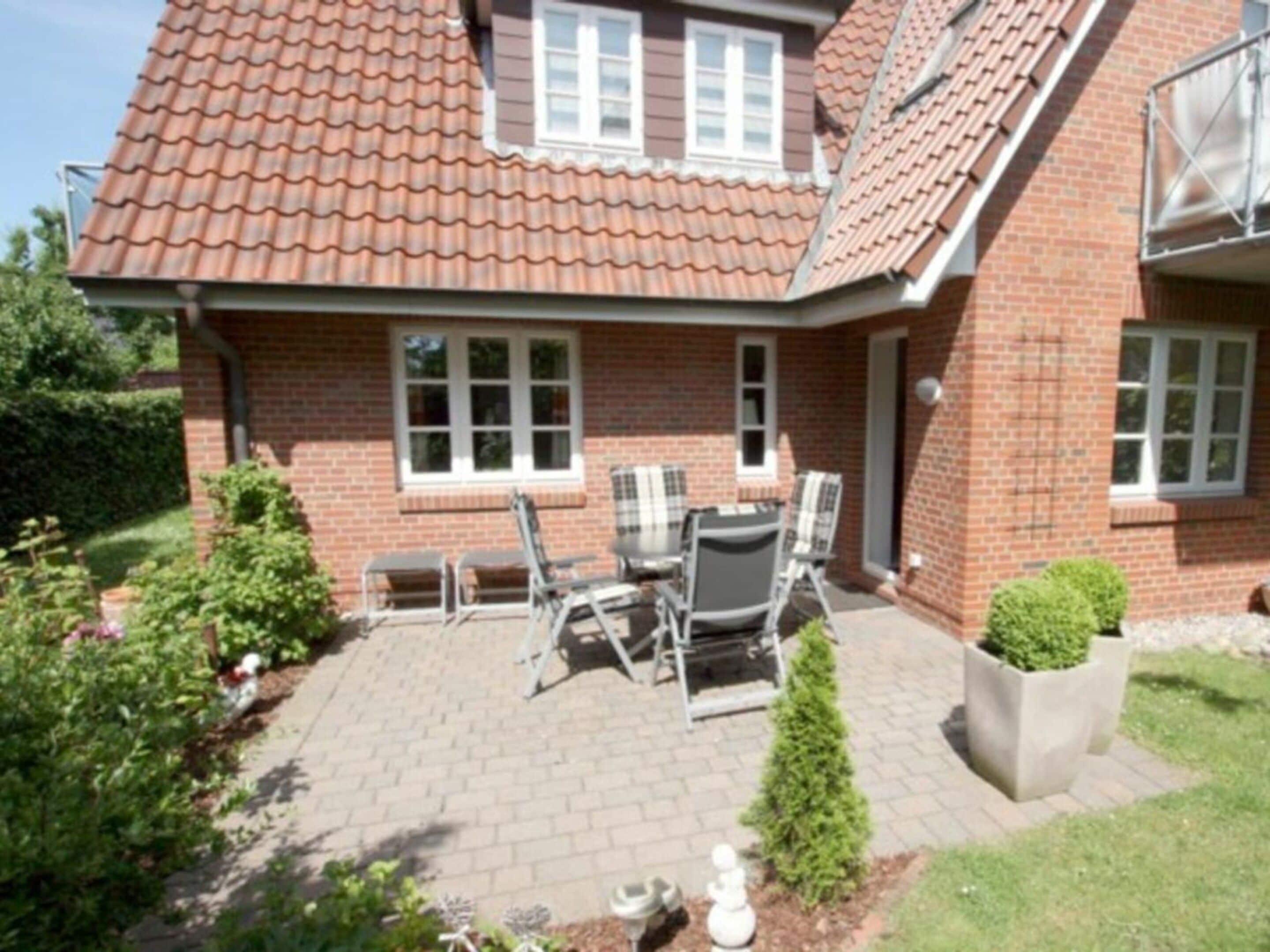 Een bakstenen huis met een pannendak, een terras met tafel en stoelen, en een kleine tuin.