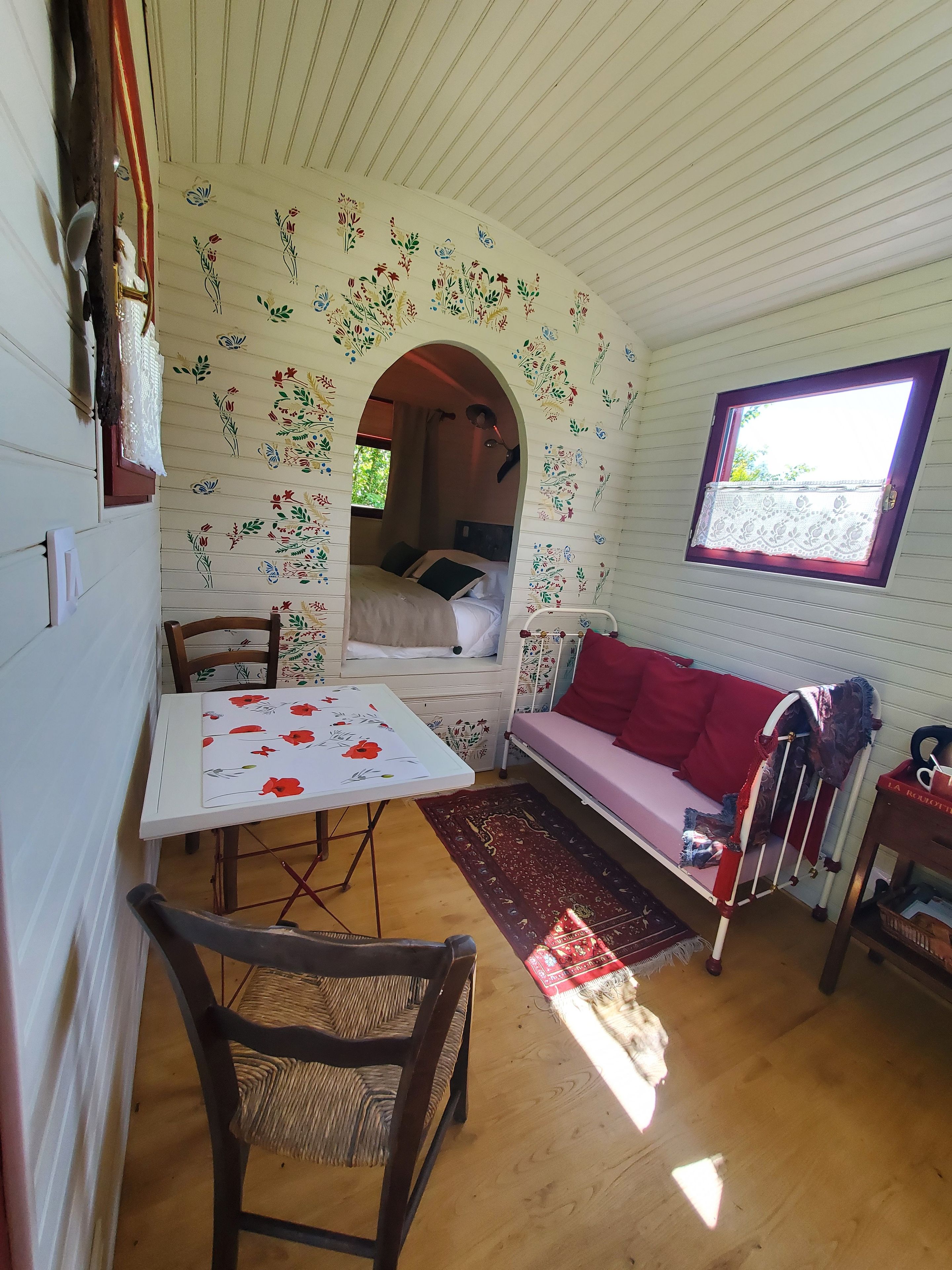 A cozy room with a bed, a red sofa, a table with a floral tablecloth, and a window with a lace curtain.