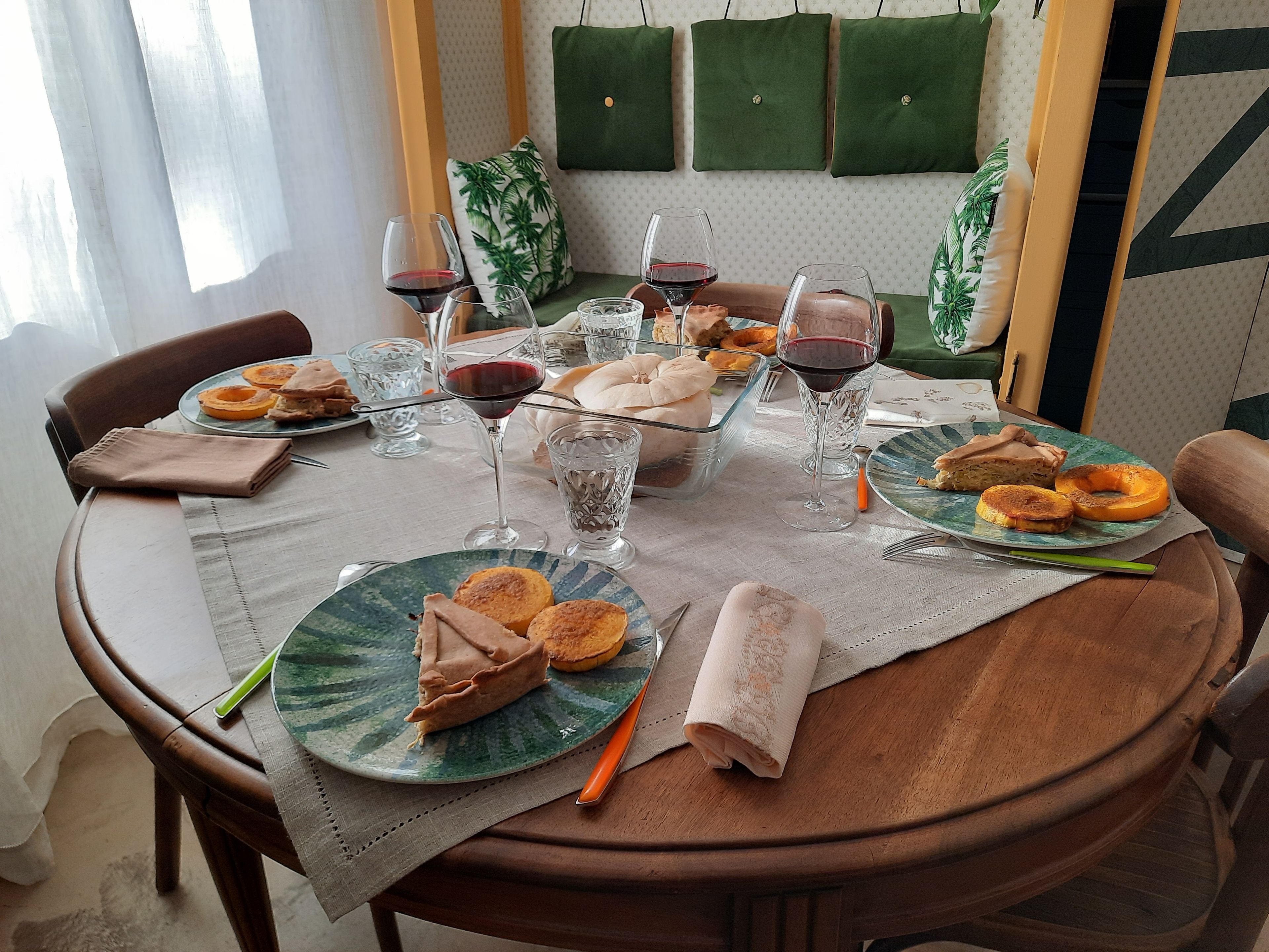 Una mesa de comedor preparada con platos de comida, copas de vino tinto y una servilleta.