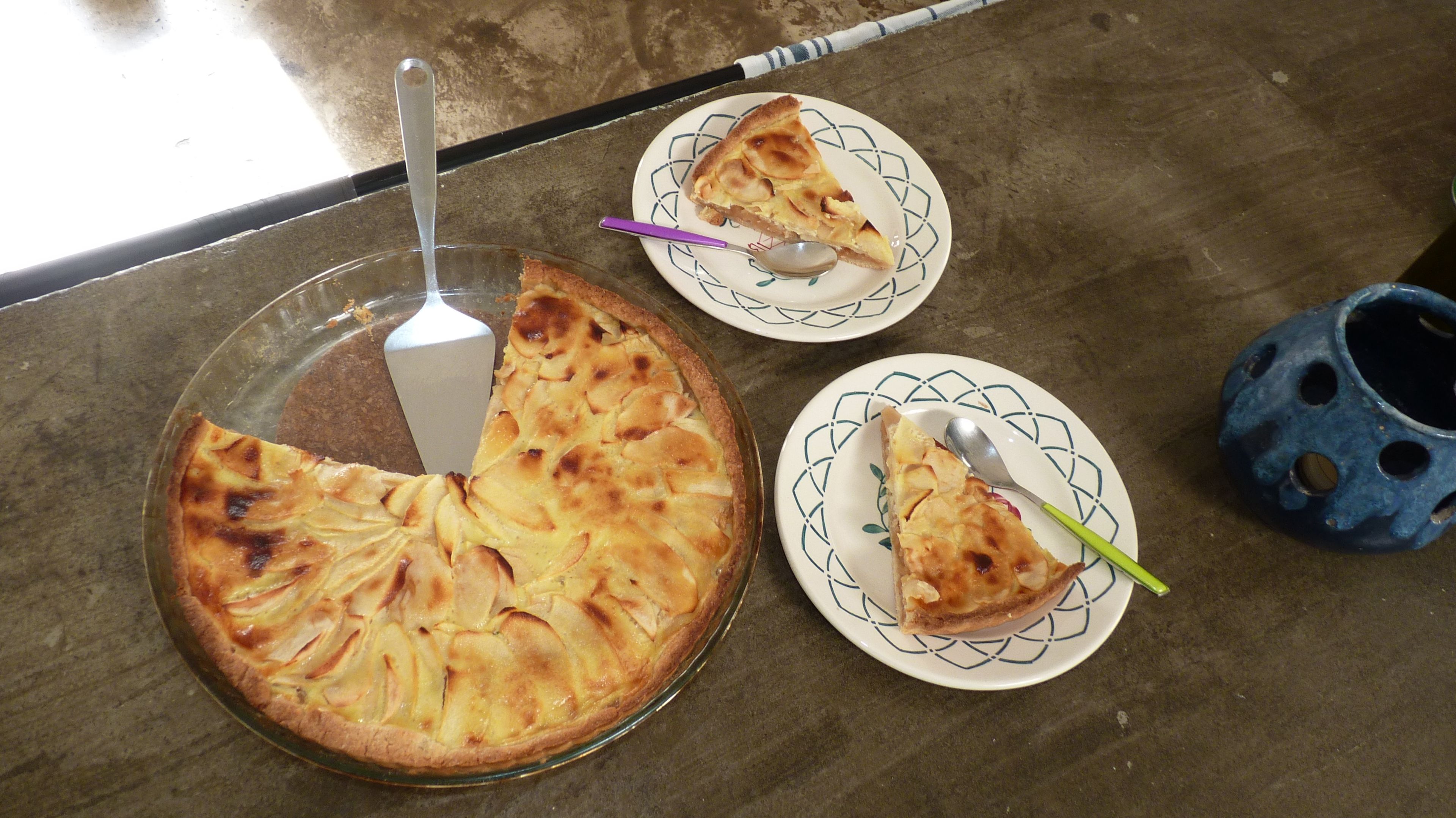 Una torta su un piatto di vetro con un cucchiaio, un'altra fetta di torta su un piatto con un cucchiaio, e una pentola di ceramica blu.