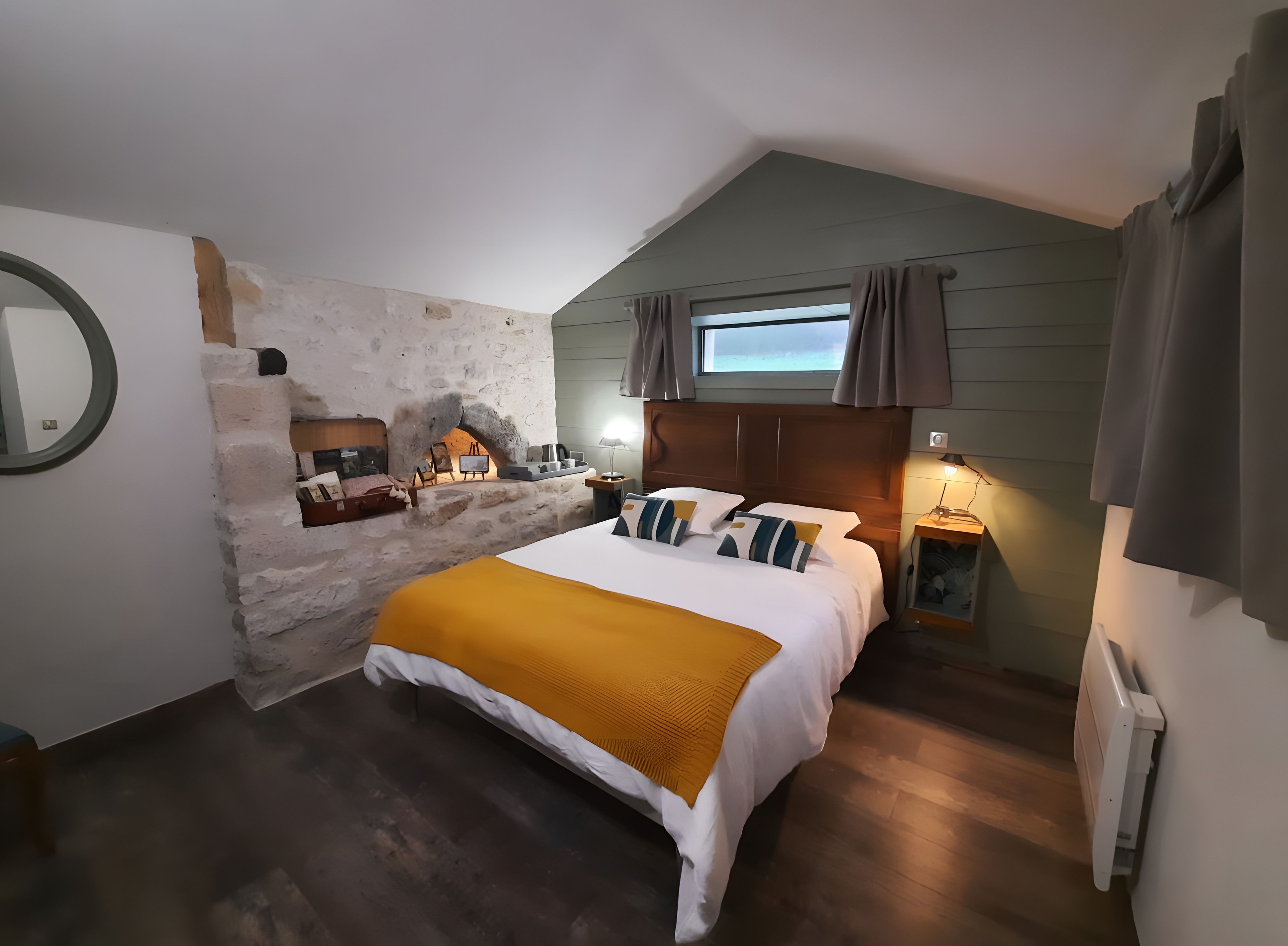 Een slaapkamer met een stenen muur, een bed met een gele plaid, een nachtkastje en een raam met gordijnen.