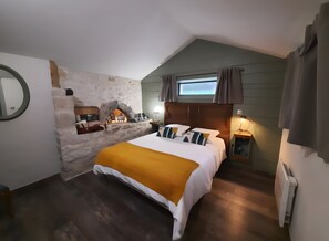 A bedroom with a stone wall, a bed with a yellow throw, a nightstand, and a window with curtains.