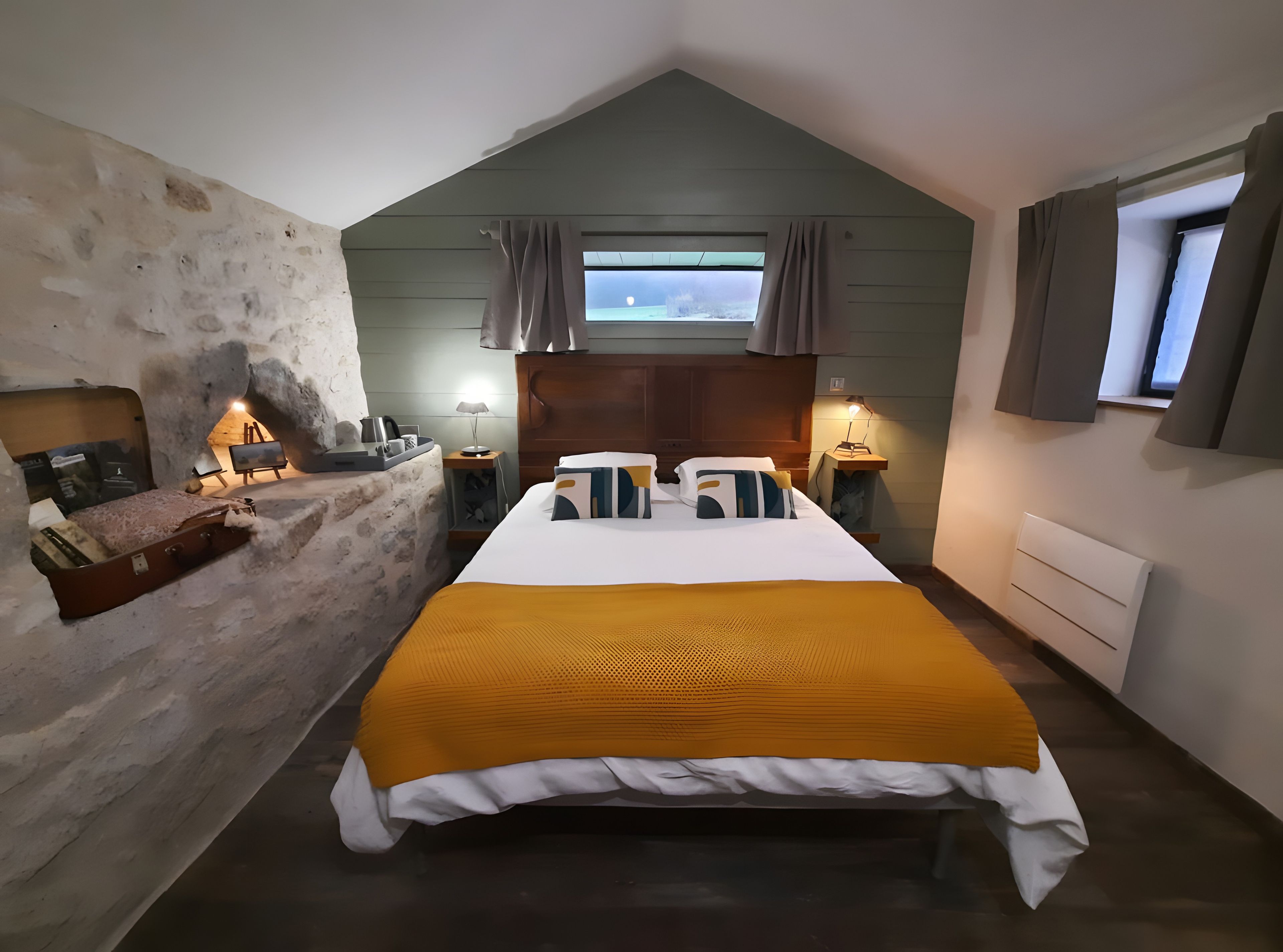 Een slaapkamer met een stenen muur, een bed met een gele deken, een houten hoofdeinde, een nachtkastje met een lamp, en een raam met gordijnen.