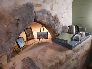 A stone fireplace with a painting on the wall, a kettle, and two mugs on a shelf.