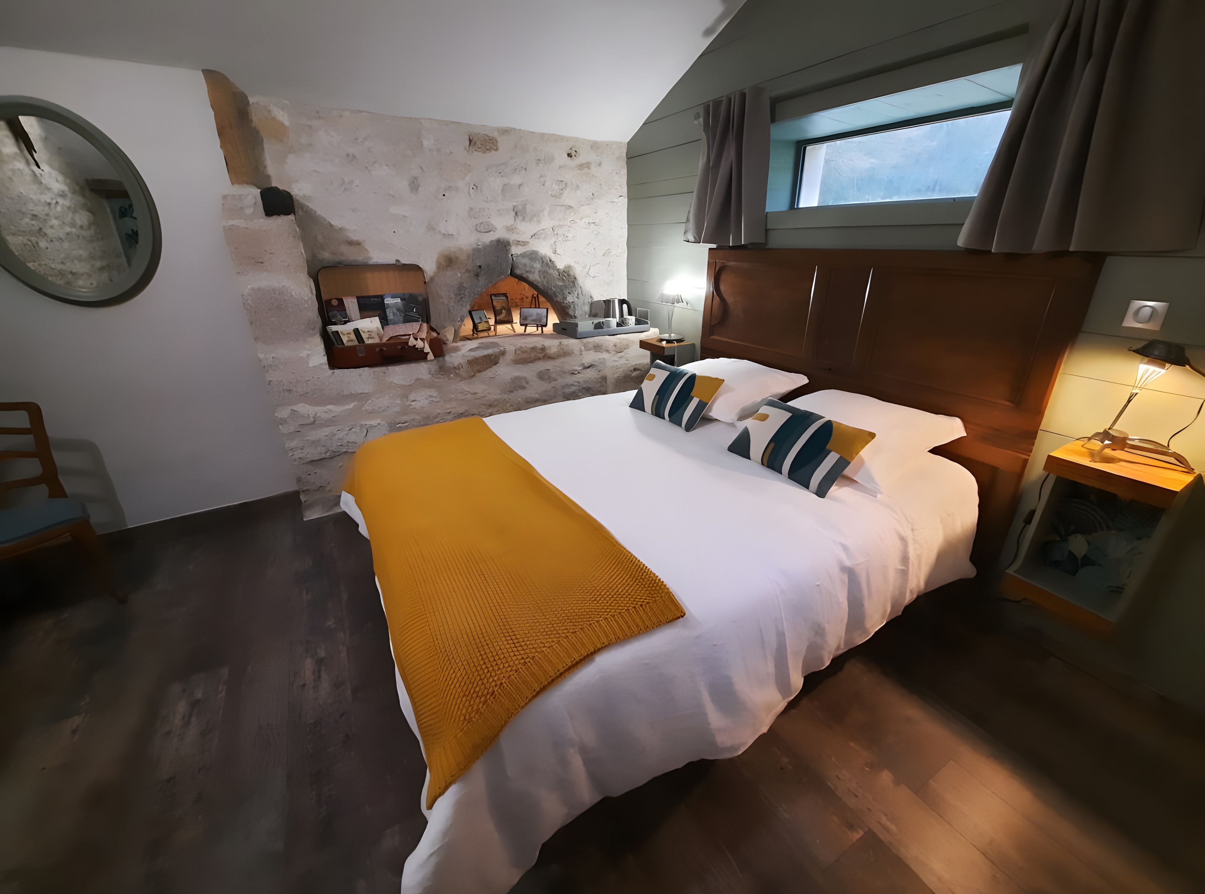 Een slaapkamer met een stenen muur, een bed met wit beddengoed en gestreepte kussens, een houten hoofdbord, een nachtkastje met een lamp en een raam met gordijnen.