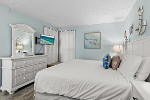A bedroom with a large bed, a dresser, a television, and a painting on the wall. - Jetty East 117a (Destin)