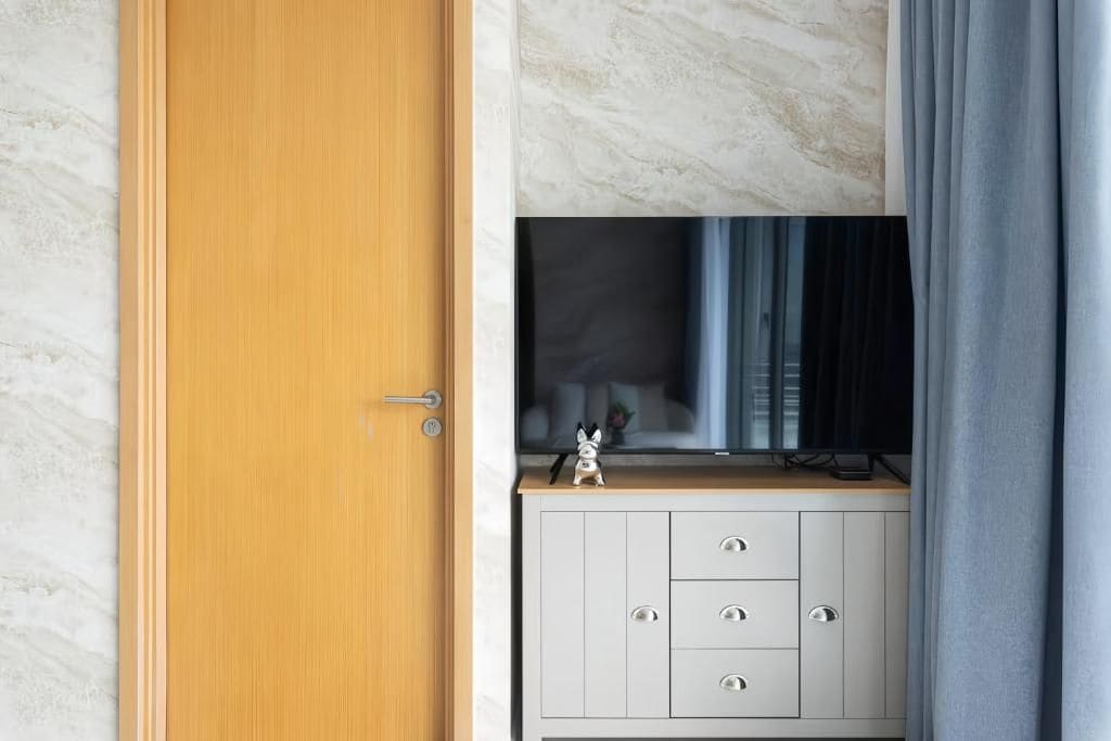 A room with a wooden door, a white cabinet with drawers, and a television on a marble wall.