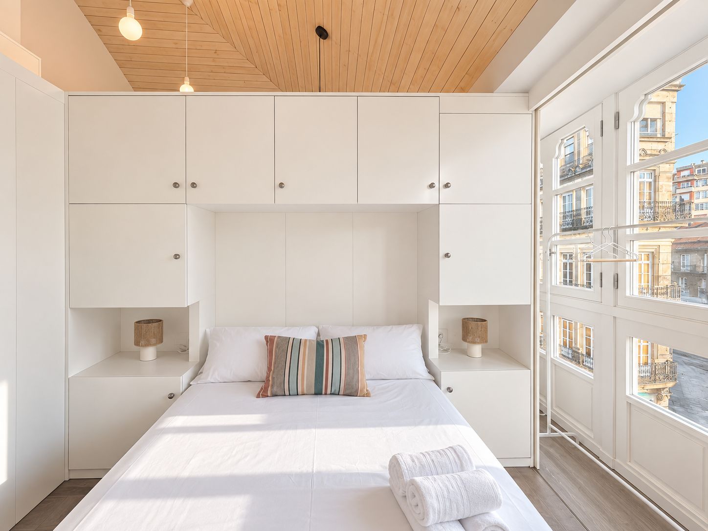 Une chambre à coucher avec un grand lit, des murs blancs, un plafond en bois et une vue sur les immeubles par la fenêtre.