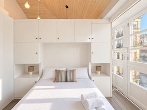Ein Schlafzimmer mit einem großen Bett, weißen Wänden, einer Holzbalkendecke und Blick auf Gebäude durch das Fenster.